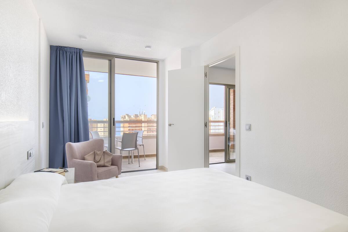 Habitación de hotel con cama, una silla, una mesa y vistas a la ciudad a través de una puerta al balcón.