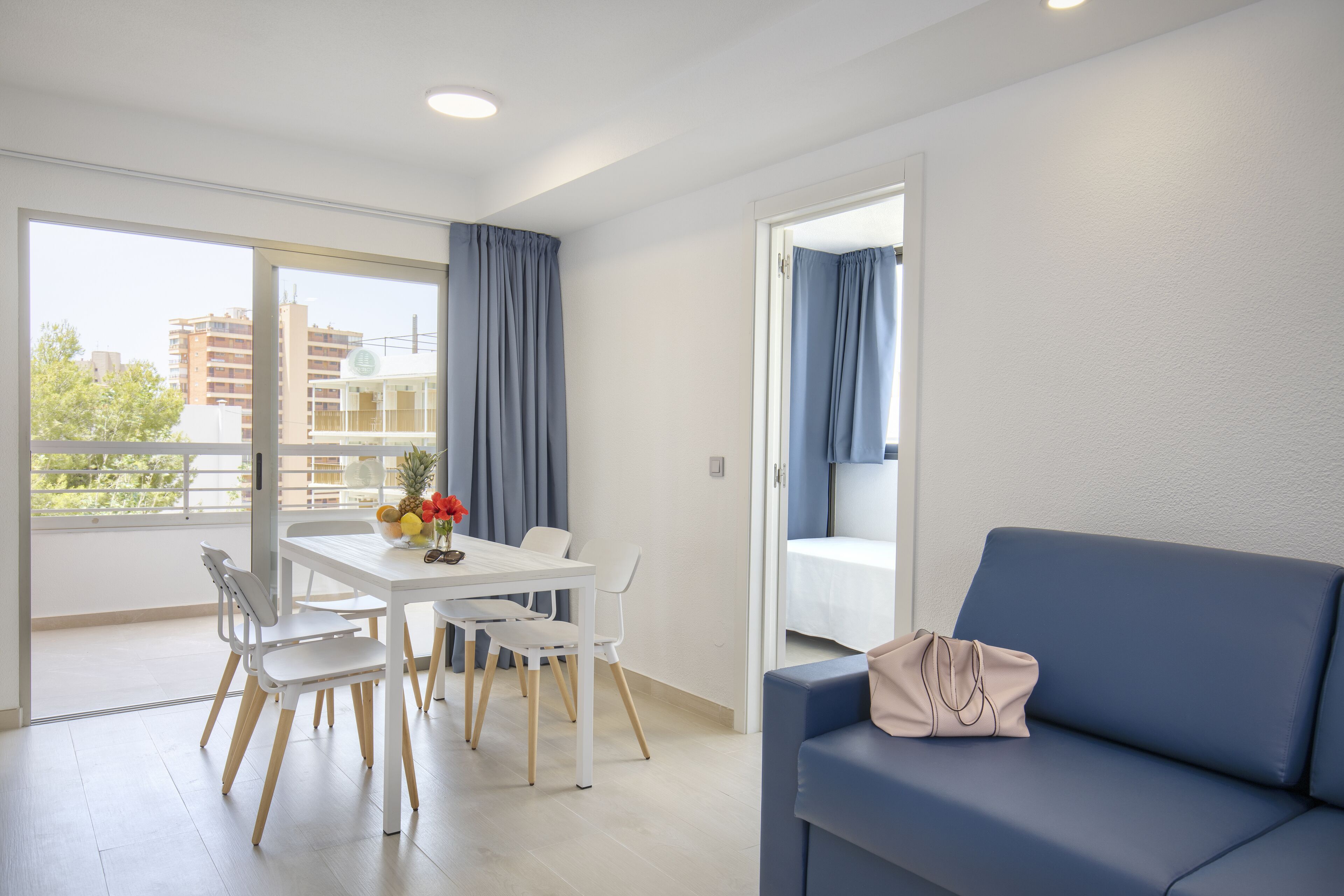 Una sala de estar moderna con una mesa blanca, cuatro sillas, un sofá azul y vistas a edificios a través de amplios ventanales.
