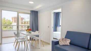 Una sala de estar moderna con una mesa blanca, cuatro sillas, un sofá azul y vistas a edificios a través de amplios ventanales.