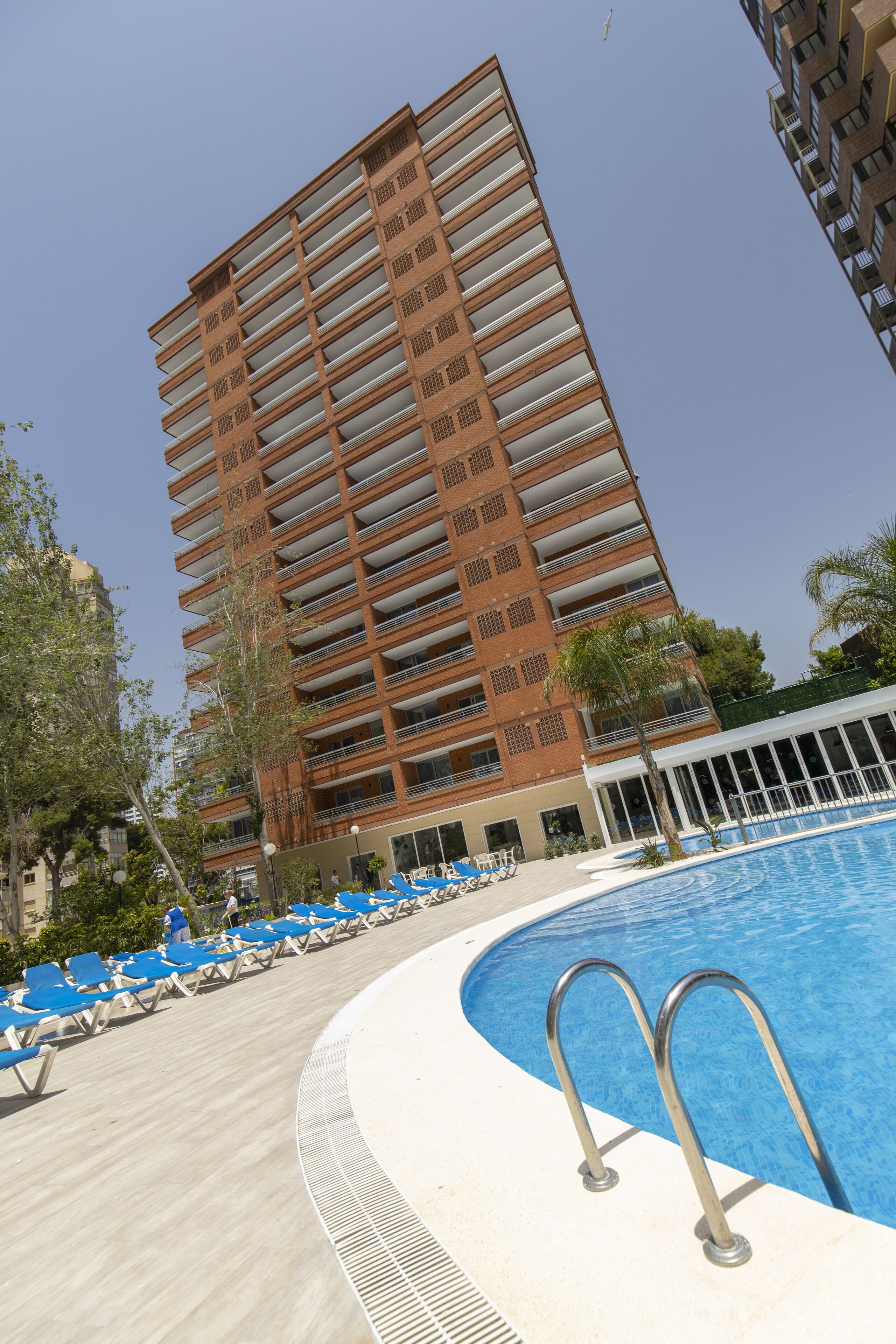 Un hotel moderno con piscina, sillas de descanso azules y cielo despejado.