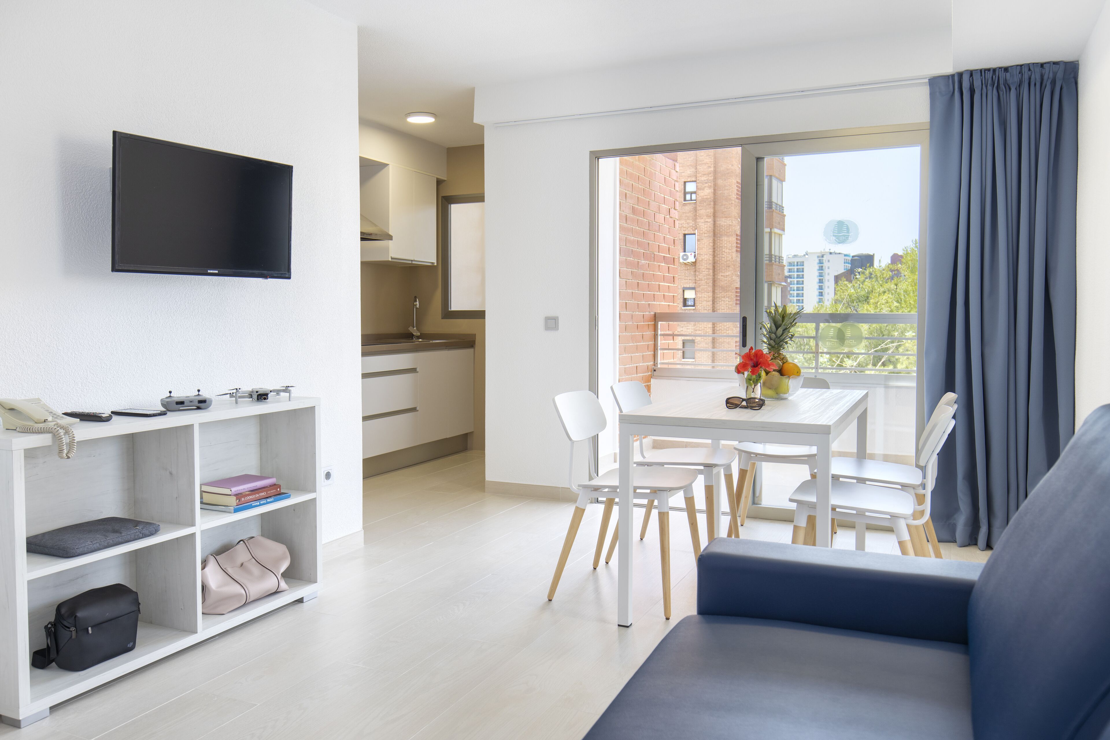 Un salon moderne avec un téléviseur à écran plat, un canapé, une table à manger avec des chaises, et un coin cuisine à l’arrière-plan.