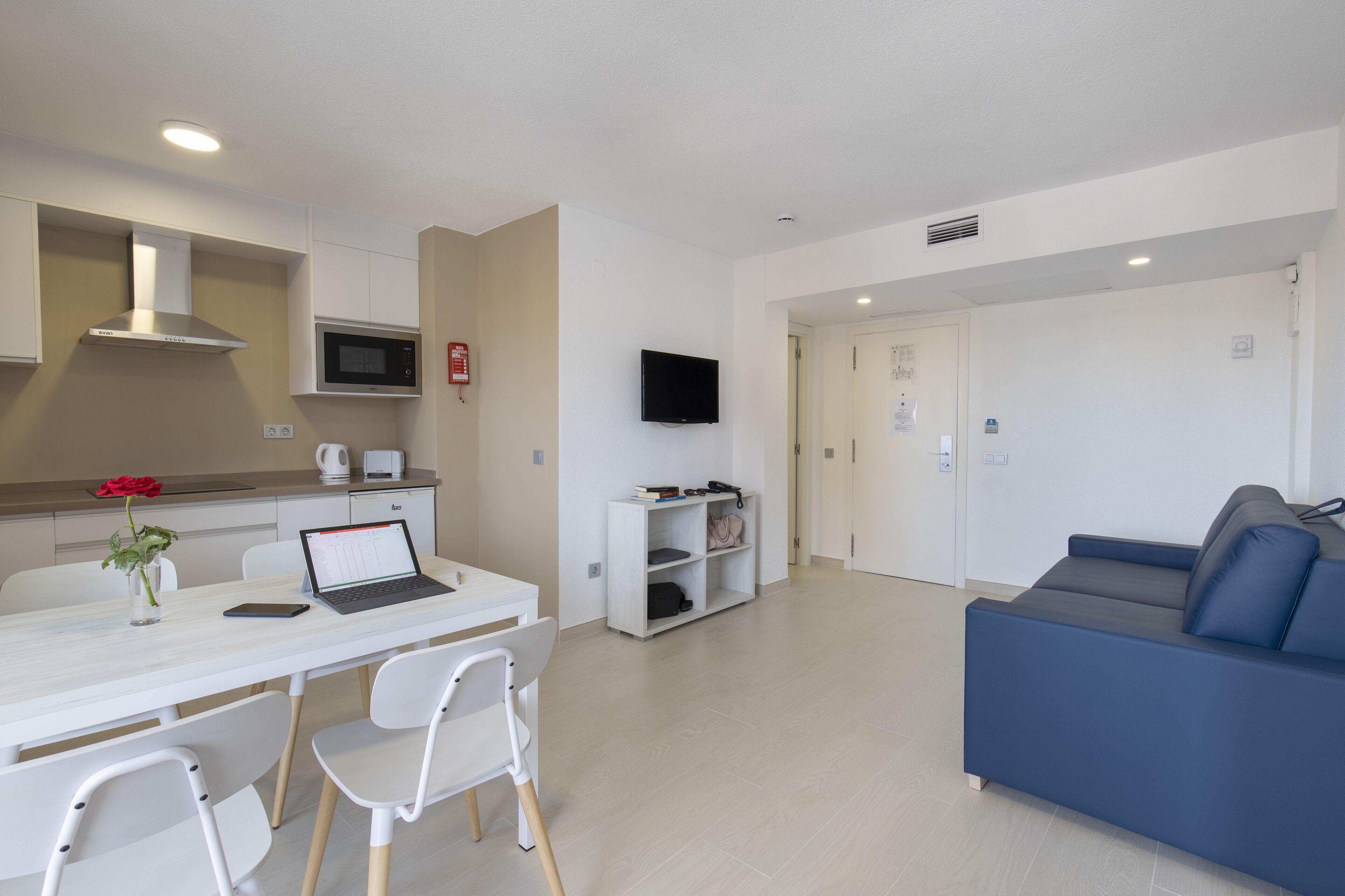 Una sala de estar moderna con un sofá azul, una mesa de comedor blanca con sillas, una zona de cocina con microondas y un televisor de pantalla plana montado en la pared.