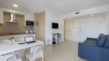 Una sala de estar moderna con un sofá azul, una mesa de comedor blanca con sillas, una zona de cocina con microondas y un televisor de pantalla plana montado en la pared.