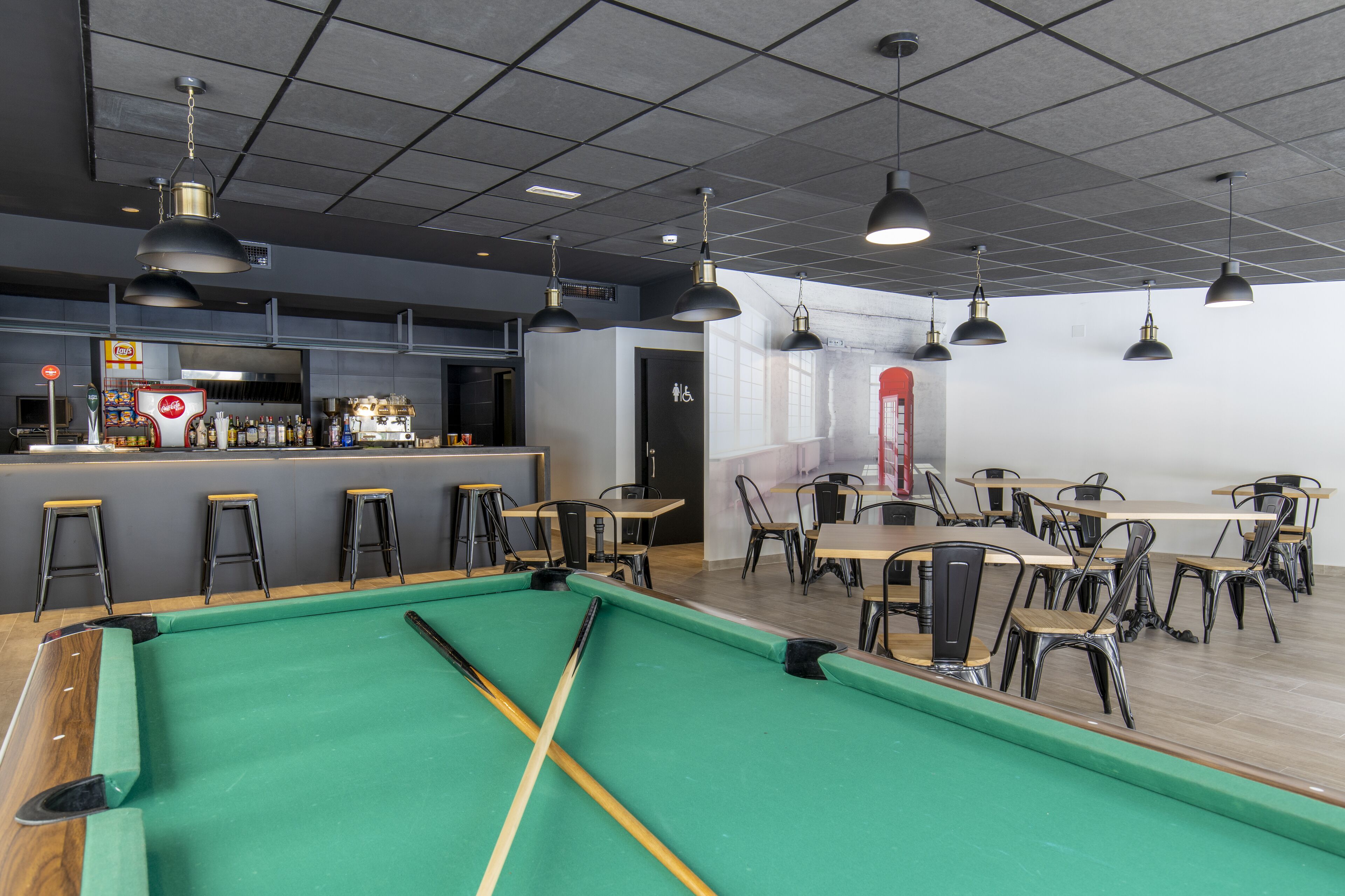 Una mesa de billar con sillas y mesas alrededor, una zona de bar con taburetes y una puerta señalizada con el símbolo de accesibilidad.
