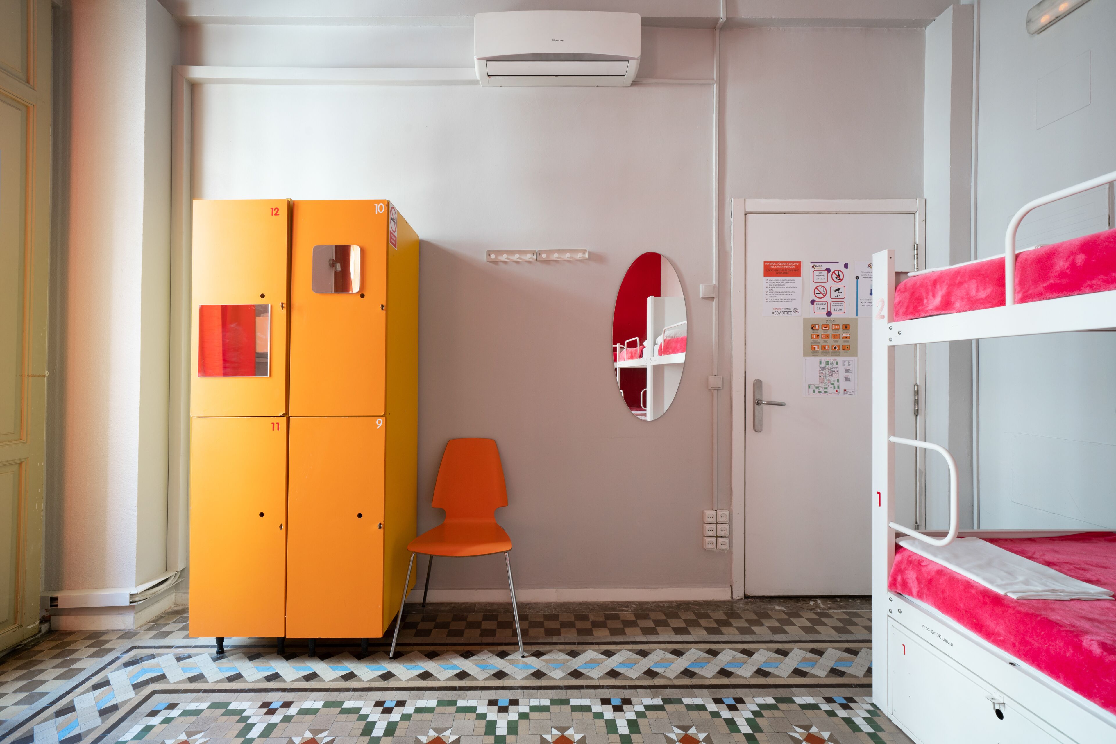 A dormitory room with bunk beds, an orange wardrobe, a checkered floor, and a door with a mirror and informational posters.