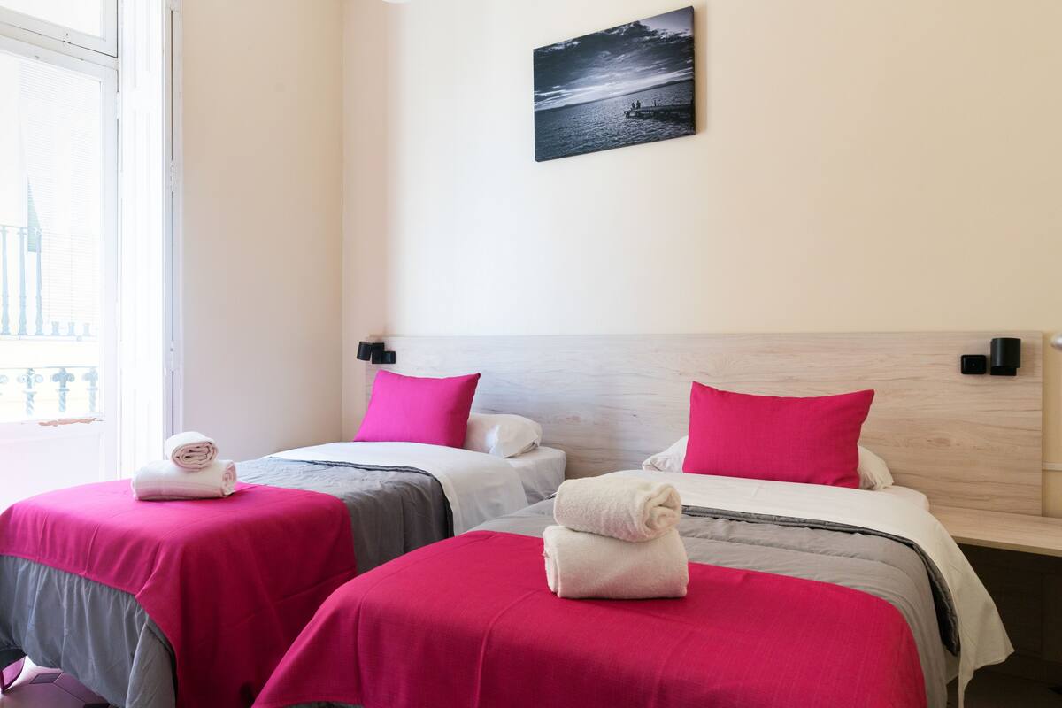 Habitación de hotel con dos camas, cada una con una colcha rosa y toallas blancas. Se ve una ventana con vistas a un edificio y un cuadro de un atardecer en la pared.