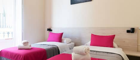 A hotel room with two beds, each with a pink bedspread and white towels. There is a window with a view of a building and a picture of a sunset on the wall.