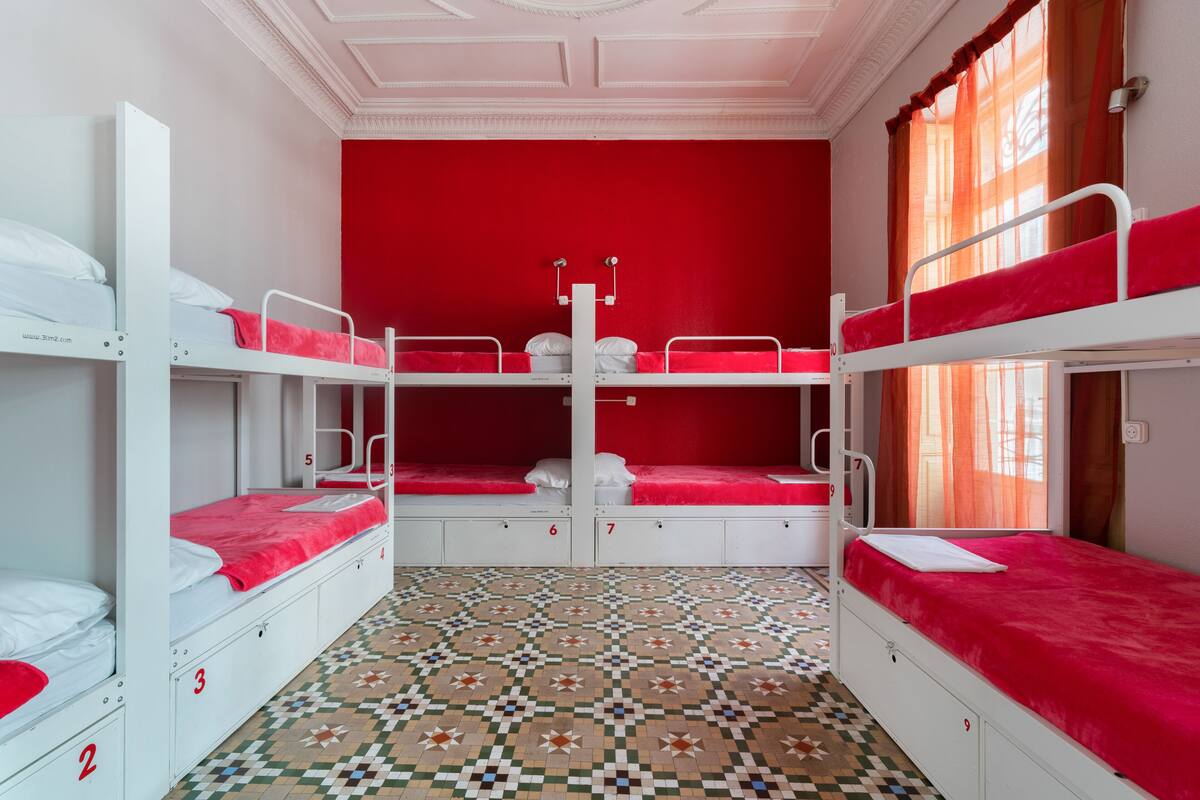 Un dormitorio con literas, camas numeradas, suelo estampado y pared roja.