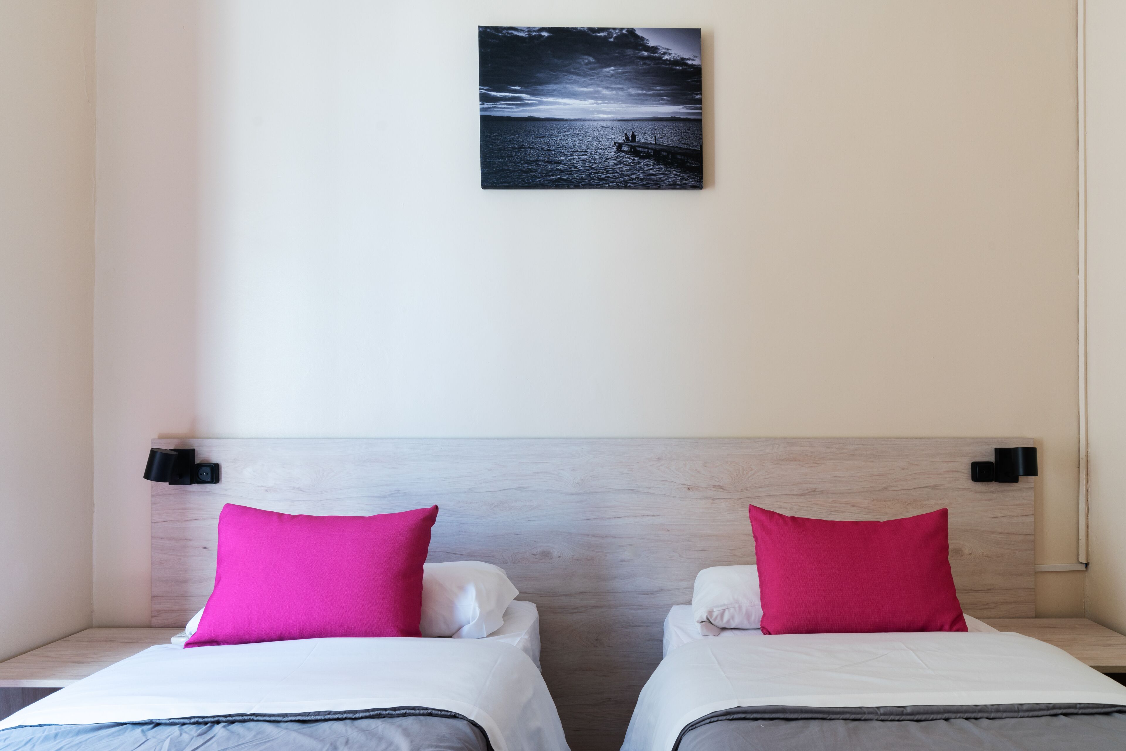 A hotel room with two beds, each with a white mattress and a bright pink pillow. There is a framed picture on the wall above the beds.