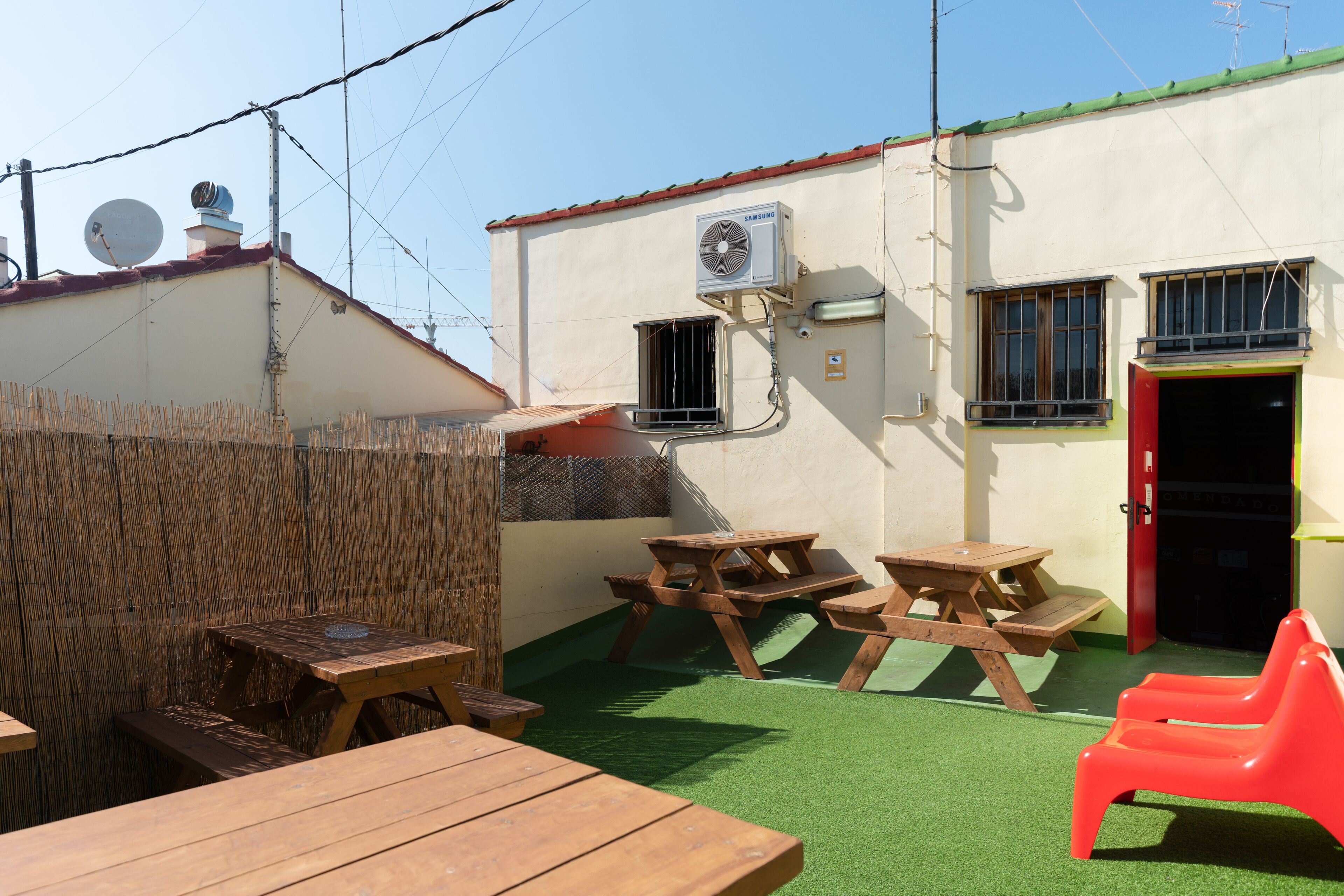 An outdoor patio area with wooden picnic tables and a red plastic chair, a wall-mounted air conditioning unit, and a door with a red frame.