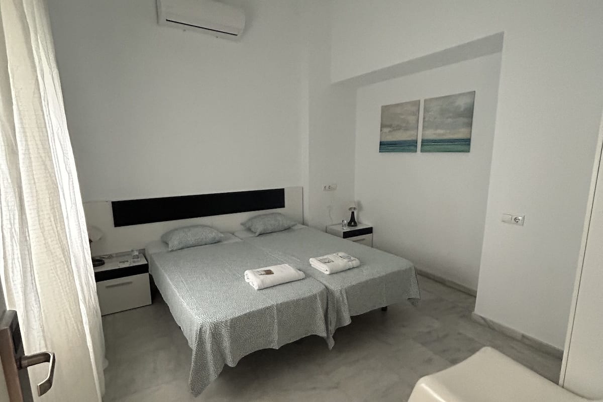 Um quarto com cama, mesas de cabeceira, ar condicionado, um banquinho e um quadro na parede.