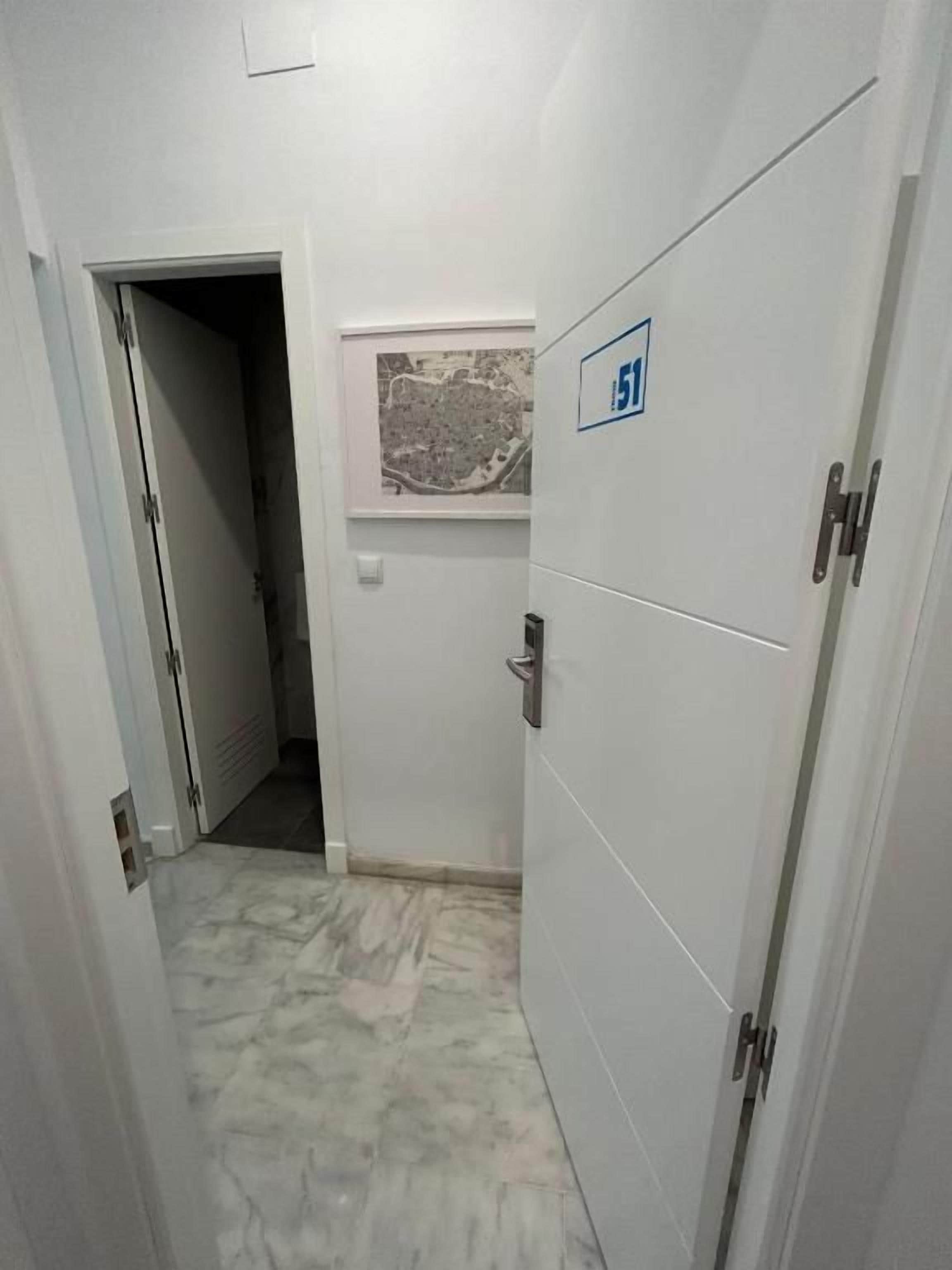 Un couloir étroit avec une porte portant un panneau indiquant qu’il est accessible aux personnes en fauteuil roulant, une fenêtre donnant sur le paysage urbain et un sol carrelé.