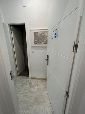 A narrow hallway with a door marked with a wheelchair accessibility sign, a window with a cityscape view, and a tiled floor. - The Spot Central Hostel (Seville)
