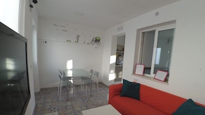 A living room with a red sofa, a TV, a dining table with chairs, and a kitchen area in the background. - The Spot Central Hostel (Seville)