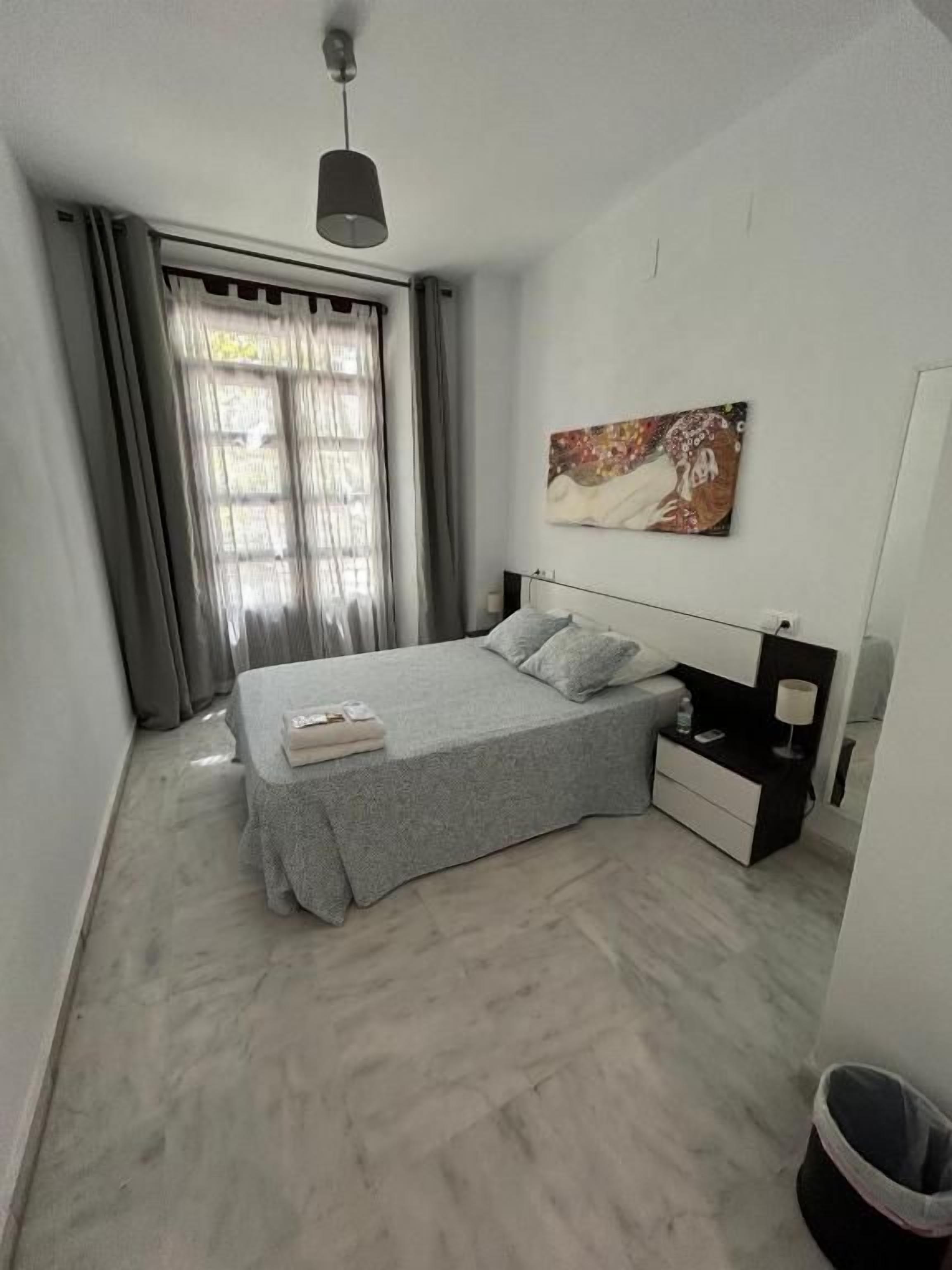 A bedroom with a bed, nightstands, a lamp, a painting, and a window with curtains.