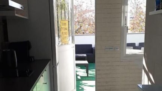 Une cuisine étroite avec des meubles blancs, un plan de travail noir et un réfrigérateur. Une porte donne accès à un espace extérieur doté d’un sol vert et d’un banc.