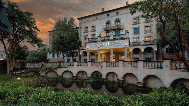 Ein historisches Gebäude mit einem 'Piazza Hotel'-Schild, das klassische Architektur, eine Brücke und ein angrenzendes Gewässer zeigt.