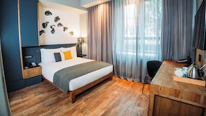 Et hotelværelse med seng, natbord, lampe, gardiner og et skrivebord med elkedel.