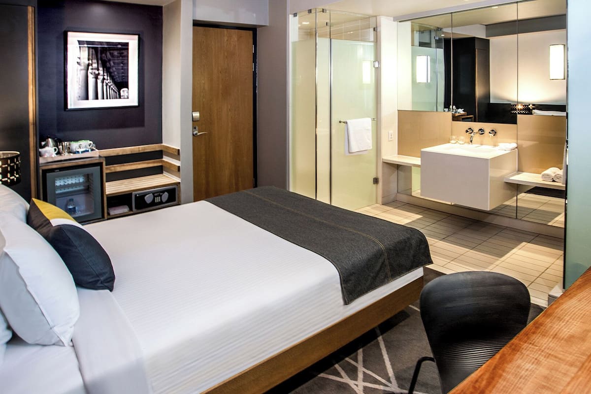 A hotel room with a bed, a desk, a mirror, a bathroom with a sink and a shower, and a small cabinet.