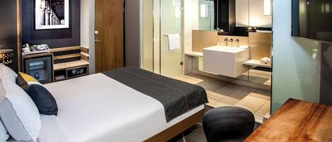 Habitación de hotel con cama, escritorio, espejo, baño con lavamanos y ducha, y un armario pequeño.