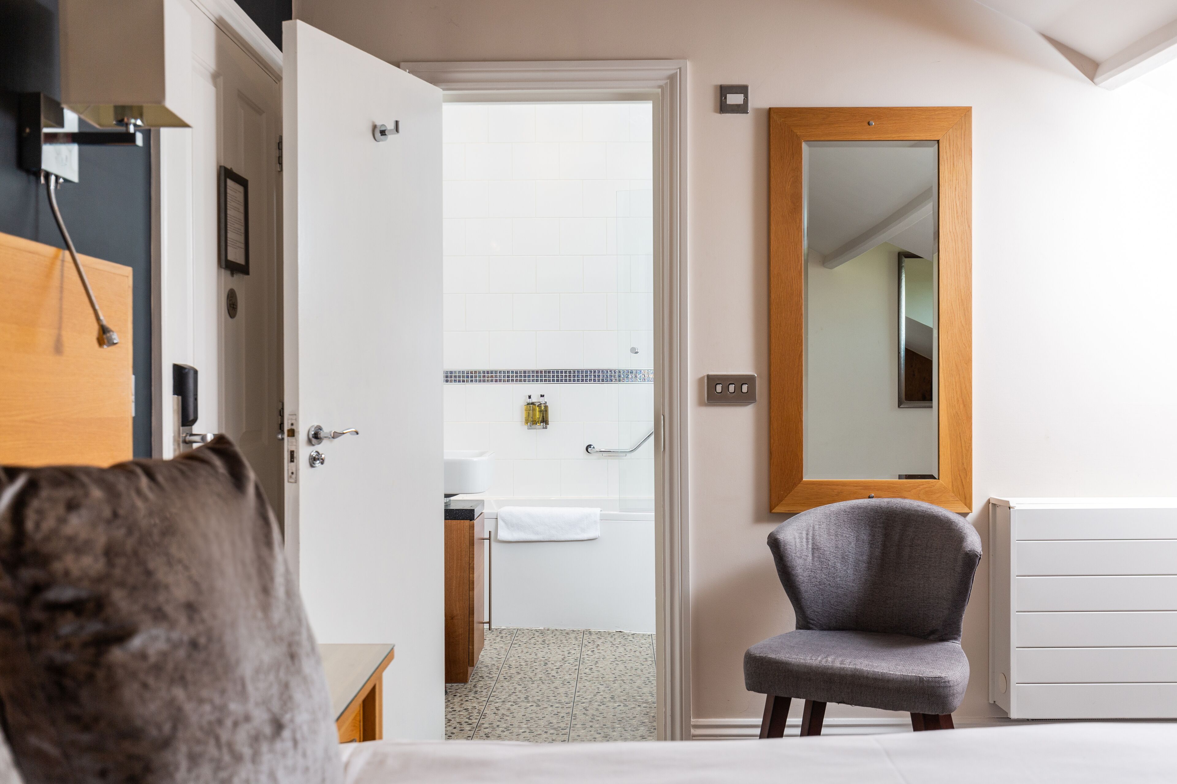A hotel room with a bed, a chair, a bathroom visible through an open door, and a large mirror on the wall.