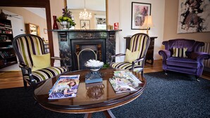 A living room with a fireplace, two armchairs, a round coffee table with magazines, and a large mirror above the fireplace. - Beaumont House (Cheltenham)