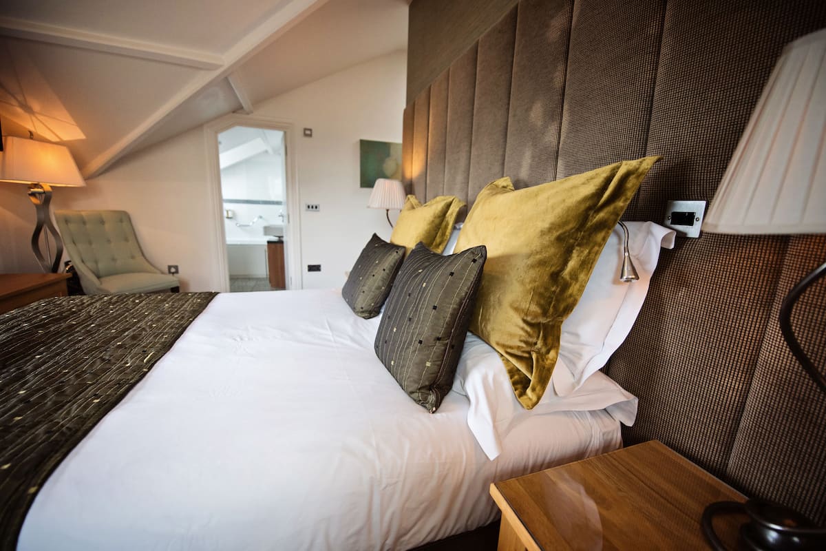 Una habitación de hotel con una cama grande, una mesita de noche de madera, una silla y una lámpara fijada a la pared.