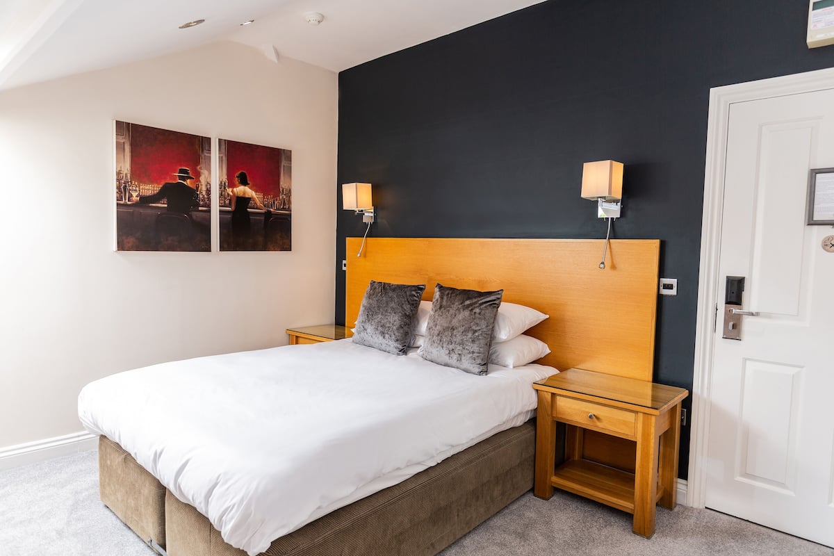 Una cama bien hecha con cabecera de madera, dos mesitas de noche con lámparas, y una puerta a la derecha.