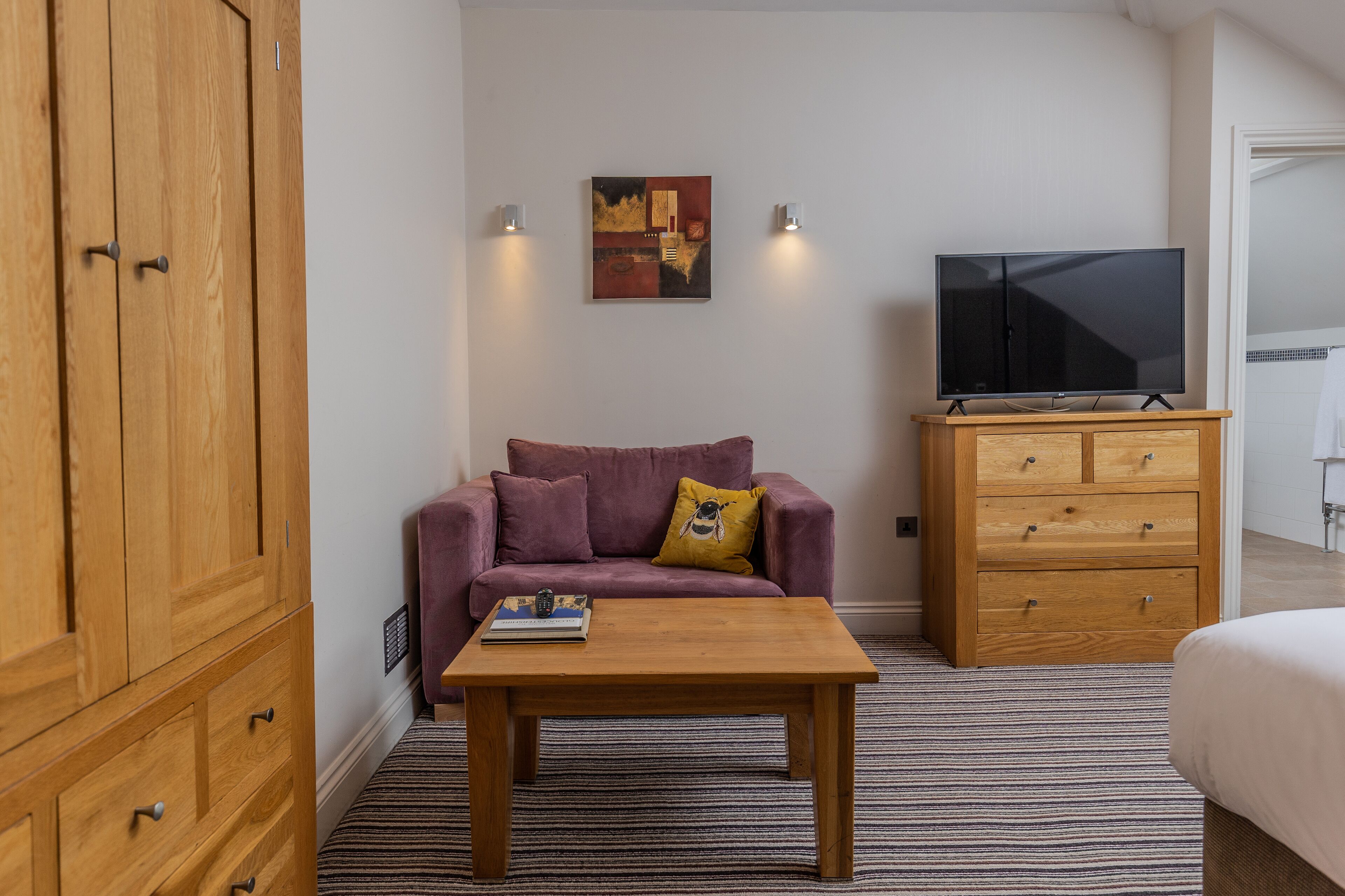 A room with a wooden wardrobe, a purple sofa with a yellow pillow, a wooden coffee table, a TV on a wooden stand, and a striped carpet.