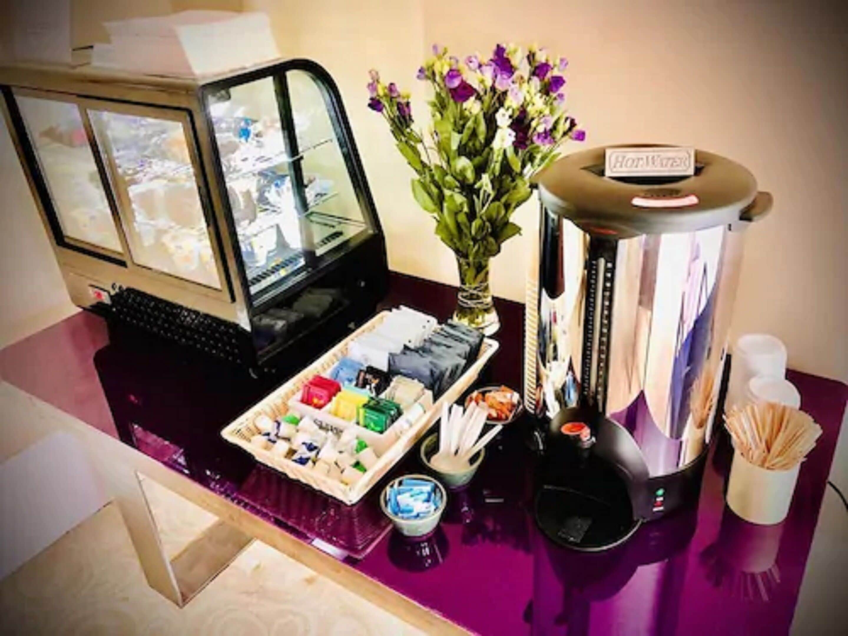 Un mostrador de recepción de hotel con una computadora, un jarrón de flores, una máquina de café y una bandeja de aperitivos.