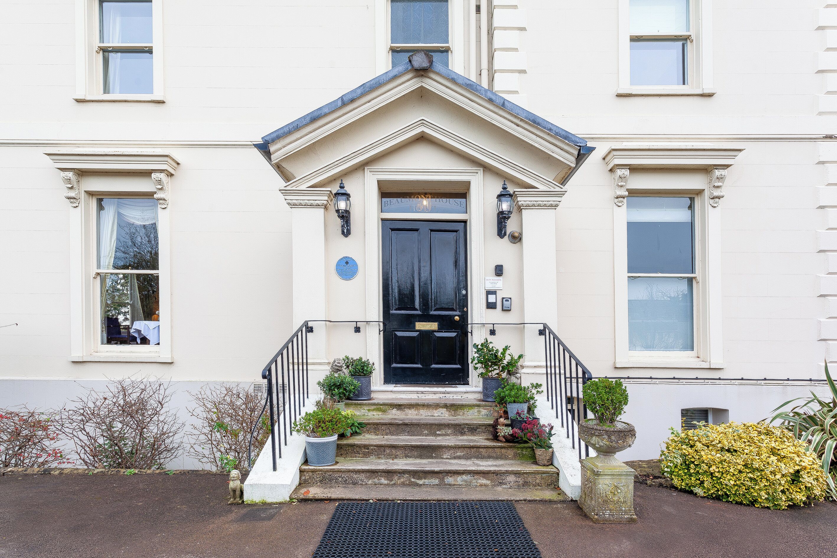 A building with a black front door, white walls, and a small porch with steps. There are potted plants on the steps and a black mat at the entrance.