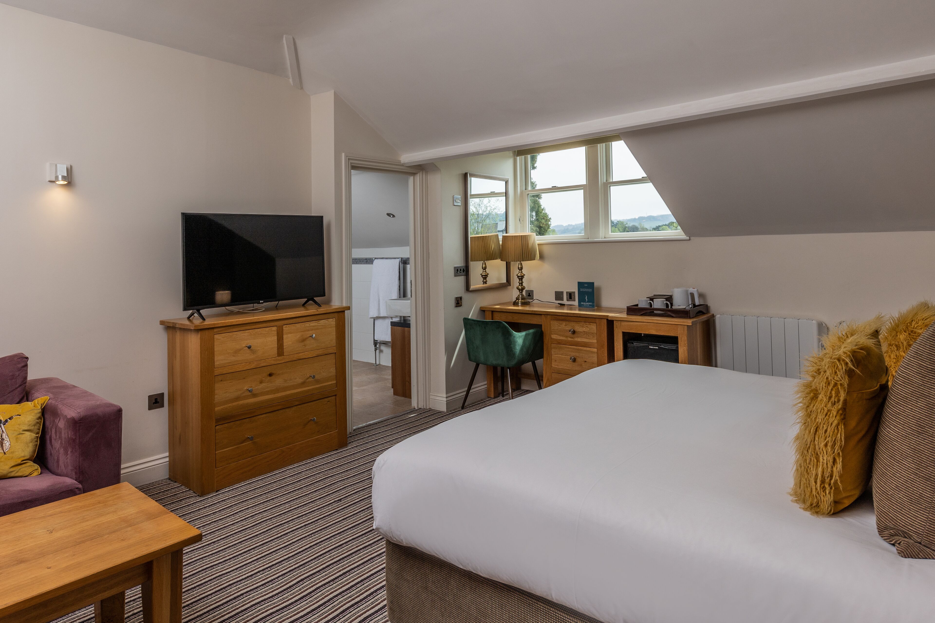 A hotel room with a large bed, a desk with a computer, a television, and a view of the outdoors through a window.