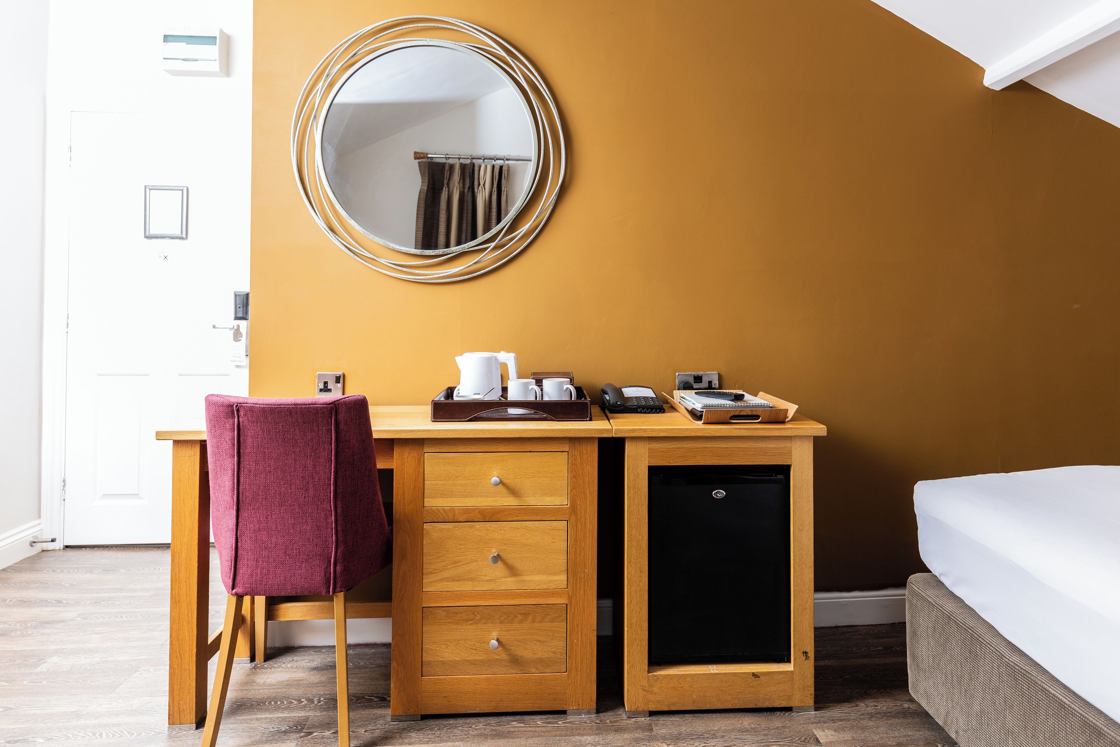 A compact hotel room with a wooden desk, a single chair, a small refrigerator, and a bed with white bedding.