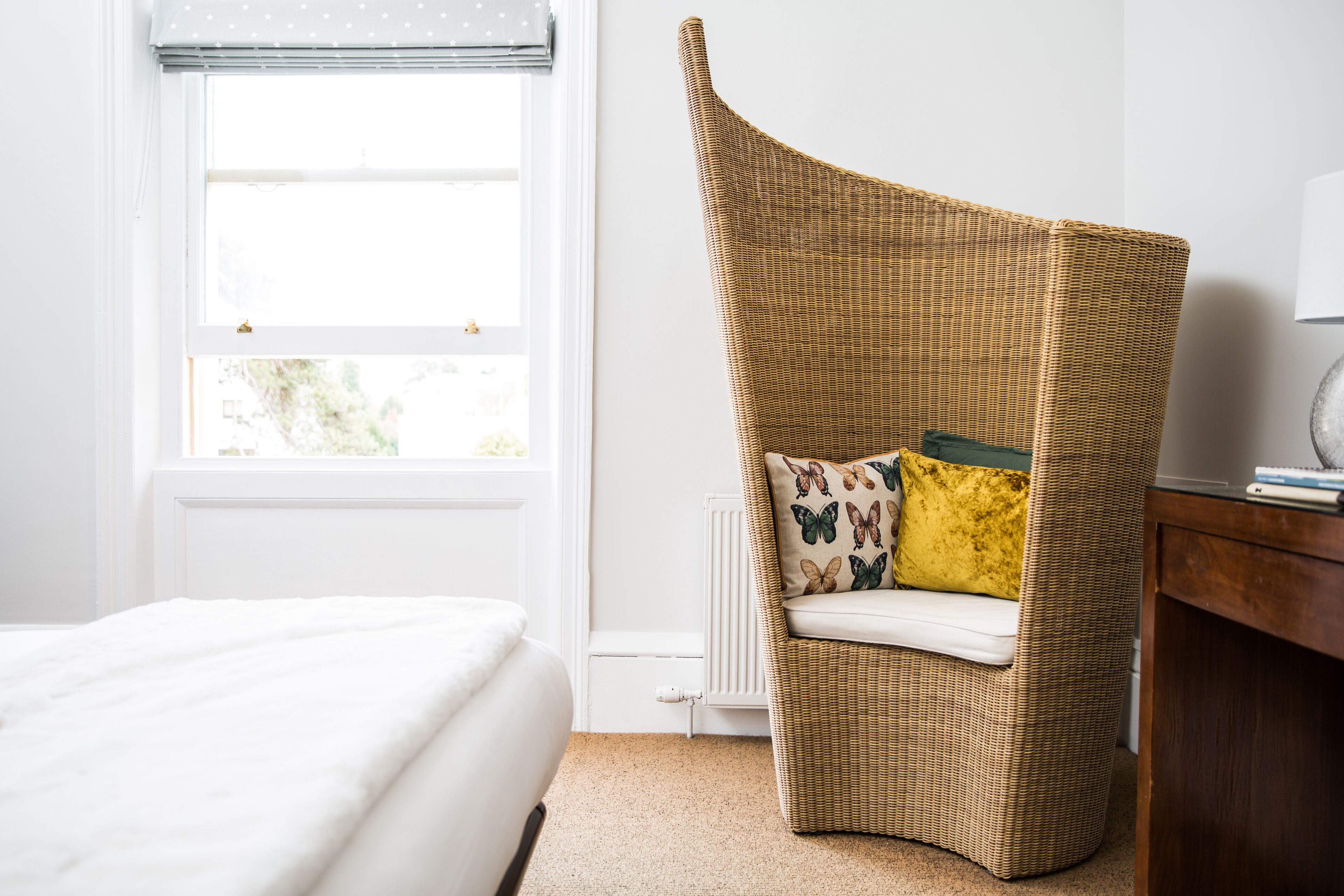 A room with a bed, a wicker chair with a cushion, a radiator, and a window with blinds.