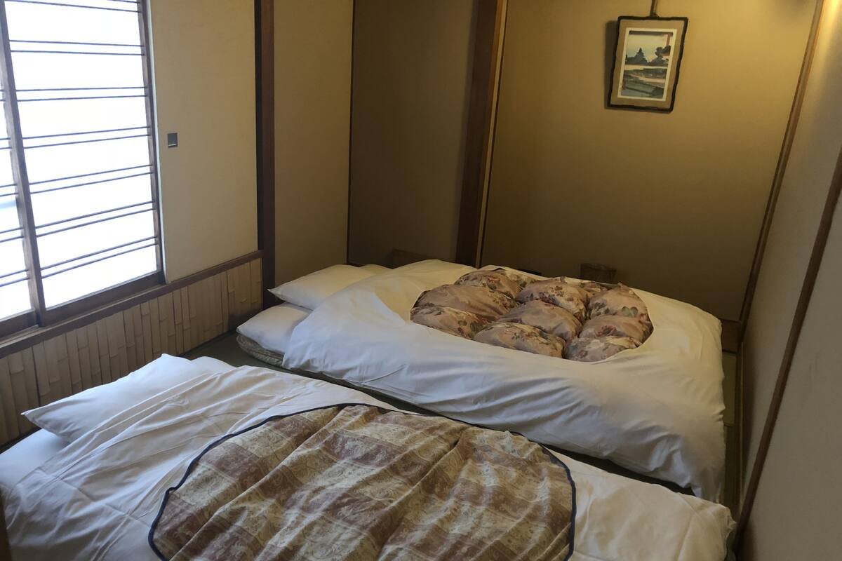 A small, compact room with two beds, one with a floral-patterned bedspread and the other with a plain white bedspread. There is a framed picture on the wall above the beds.