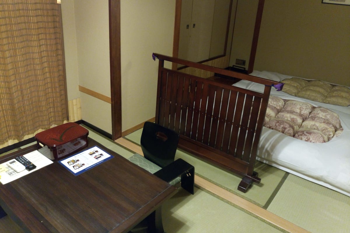A hotel room with a wooden desk, a chair, a bed with pillows, a nightstand, and a mirror.