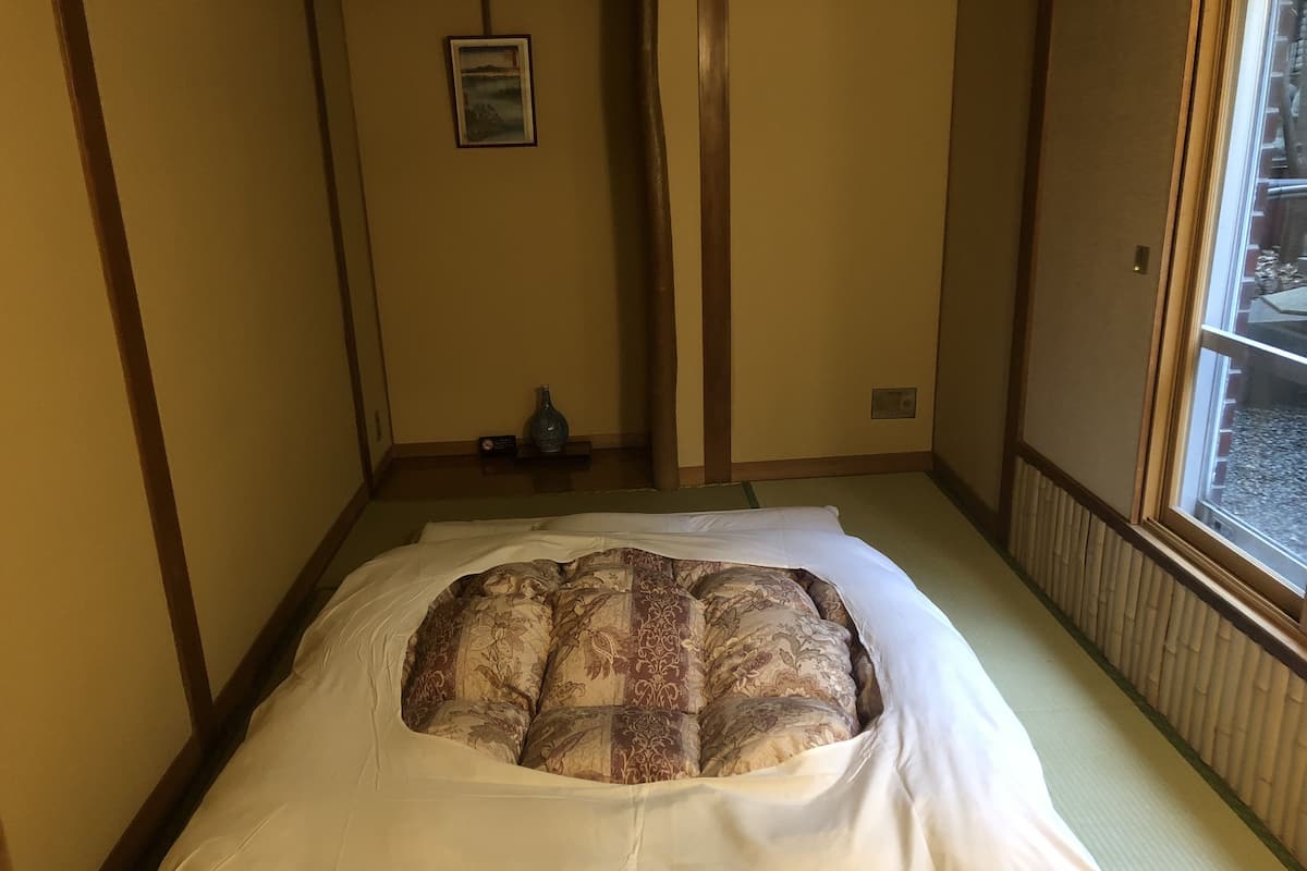 A traditional Japanese-style room with tatami flooring, a low bed with a patterned bedspread, a small vase, and a framed picture on the wall.