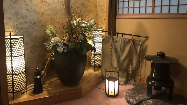 Une pièce de style japonais traditionnel, avec un sol recouvert de tatamis, une grande plante en pot, deux lanternes et une petite table agrémentée d’un objet décoratif.