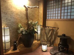 Una habitación japonesa tradicional con suelo de tatami, una planta grande en maceta, dos faroles y una mesita con un objeto decorativo.