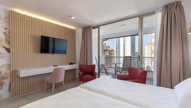 Habitación de hotel con cama, escritorio con silla, televisor montado en un panel de madera y balcón con vista a edificios.