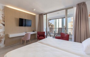 Habitación de hotel con cama, escritorio con silla, televisor montado en un panel de madera y balcón con vista a edificios.