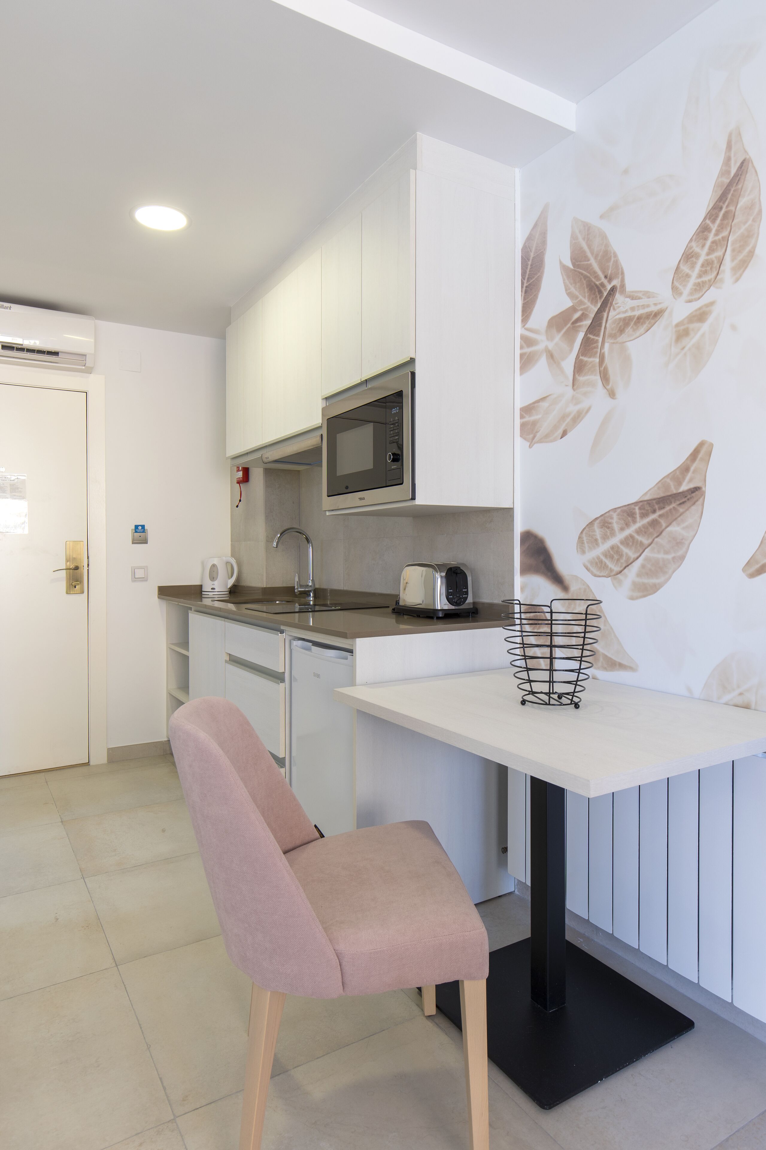 Una kitchenette compacta con microondas, tostadora y tetera, una silla pequeña y una pared con diseño de hojas.