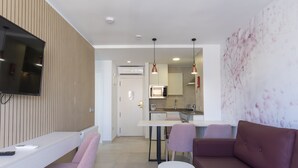Una sala de estar moderna con un televisor de pantalla plana, una zona de comedor con una mesa y sillas blancas, y una cocina al fondo.