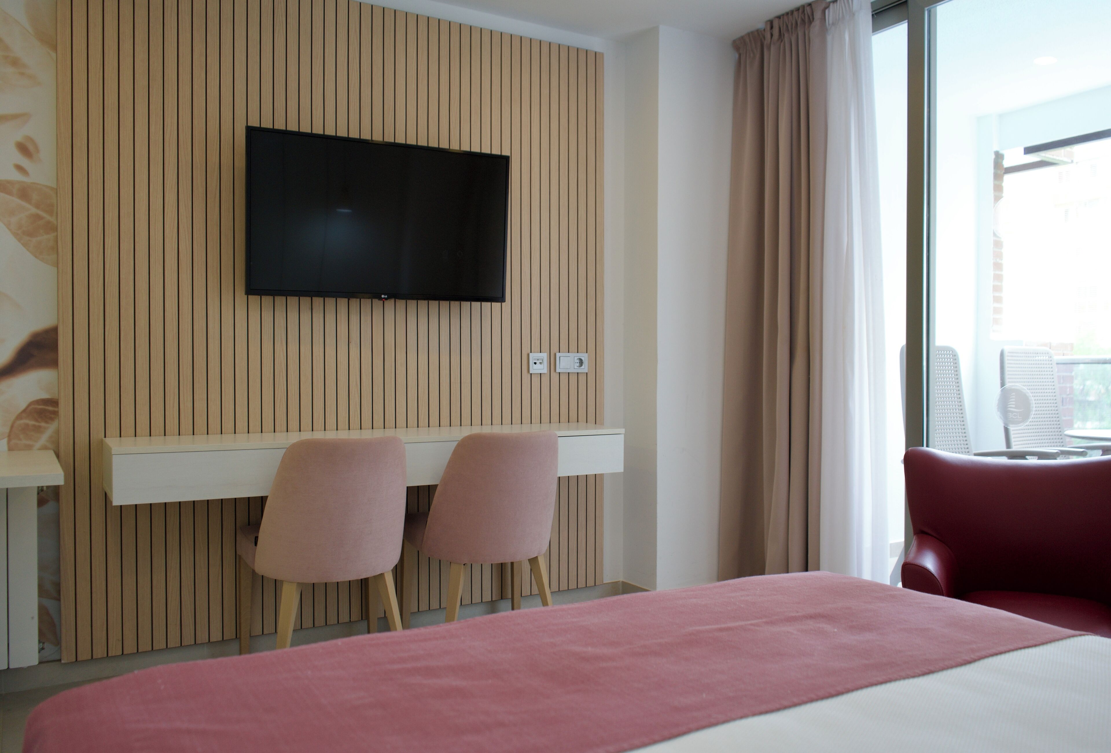 A hotel room with a bed, a TV mounted on a wooden panel wall, two pink chairs, and a red chair.