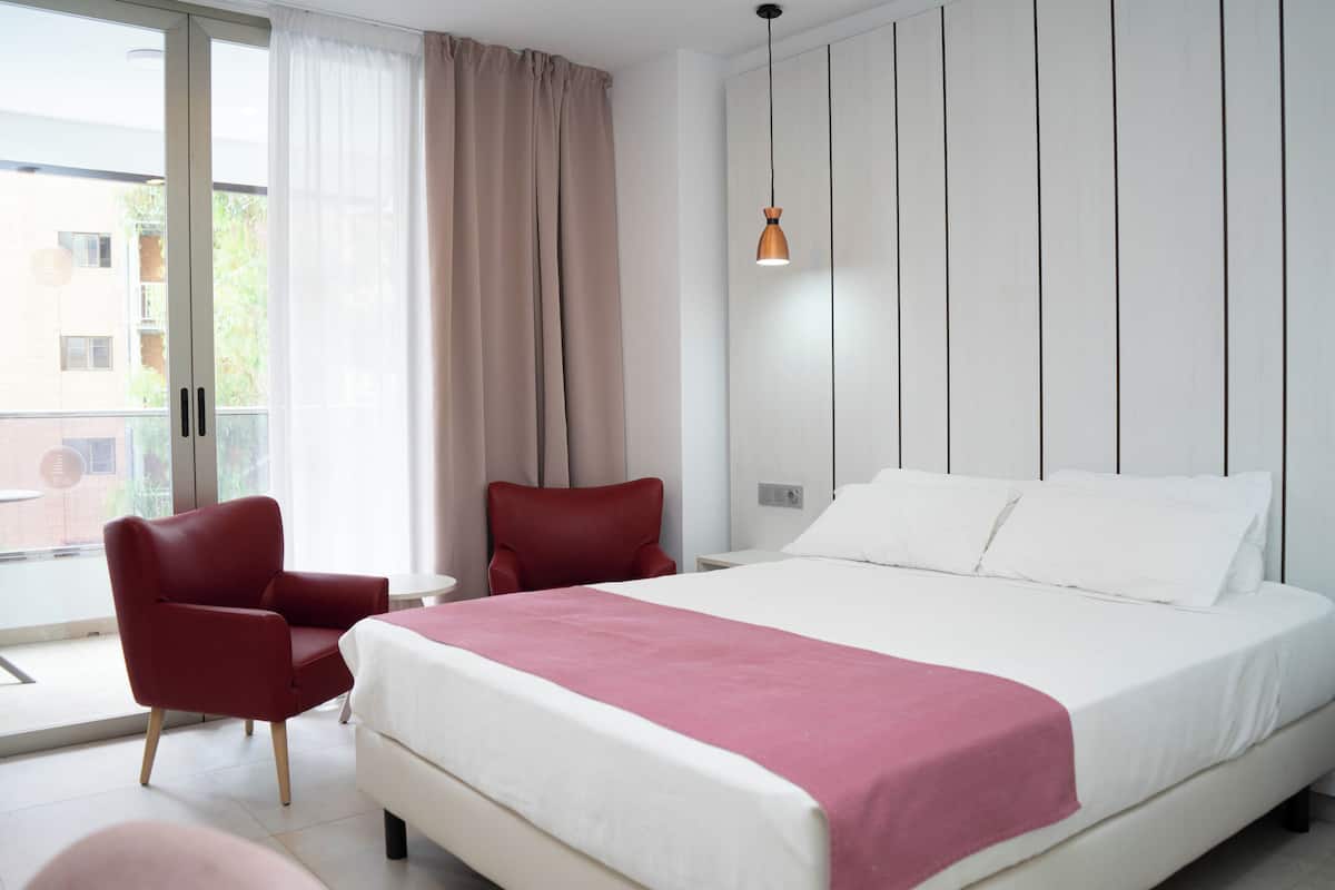 Una habitación de hotel moderna con una cama grande, un sillón rojo, una mesita y una ventana con cortinas.
