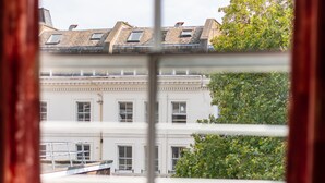 A view through a window with white blinds, revealing a building with multiple stories, a balcony, and a tree in the foreground. - Dolphin Inn (London)