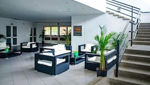 Un hall d’entrée moderne avec un escalier, un espace détente avec des canapés et des tables, et un panneau affichant un logo.