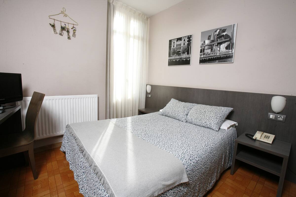 Hotellihuone, jossa on sänky, työpöytä, tuoli, patteri, puhelin ja kaksi mustavalkoista valokuvaa seinällä.