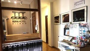 Uma área de recepção com balcão de madeira, uma placa com 'RIPAHOTEL BILBAANO' e uma mesa exibindo vários folhetos e revistas.