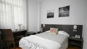 Quarto de hotel com cama, escrivaninha, cadeira e mesa de cabeceira com telefone.
